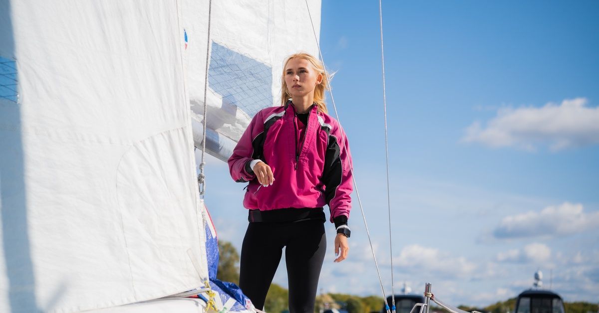 A young blonde woman in a pink jacket standing on a sailboat.