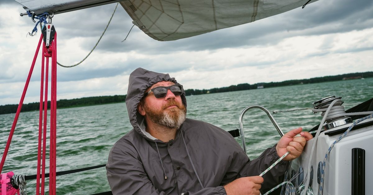 A man wearing a hooded jacket and sunglasses using a slim green-and-white rope to control the sails on a sailboat.