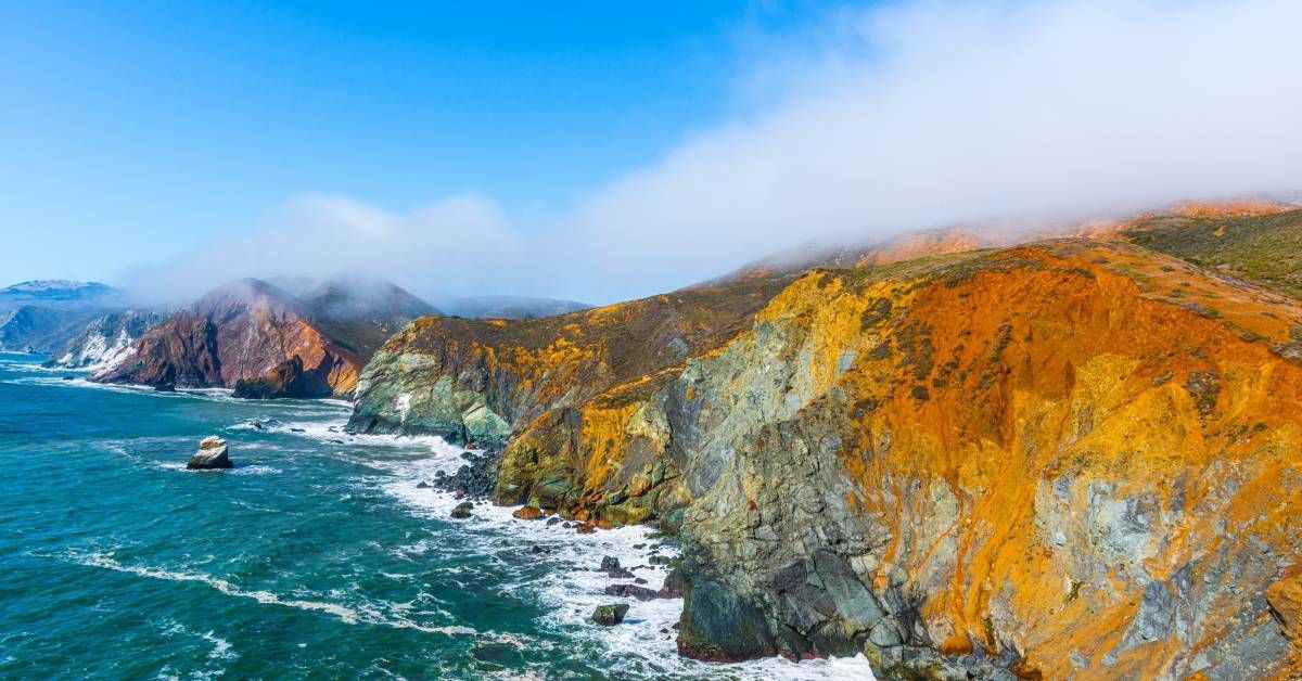 An orange-oxidized coastline overlooking turquoise water with crashing white waves and a large cloud.