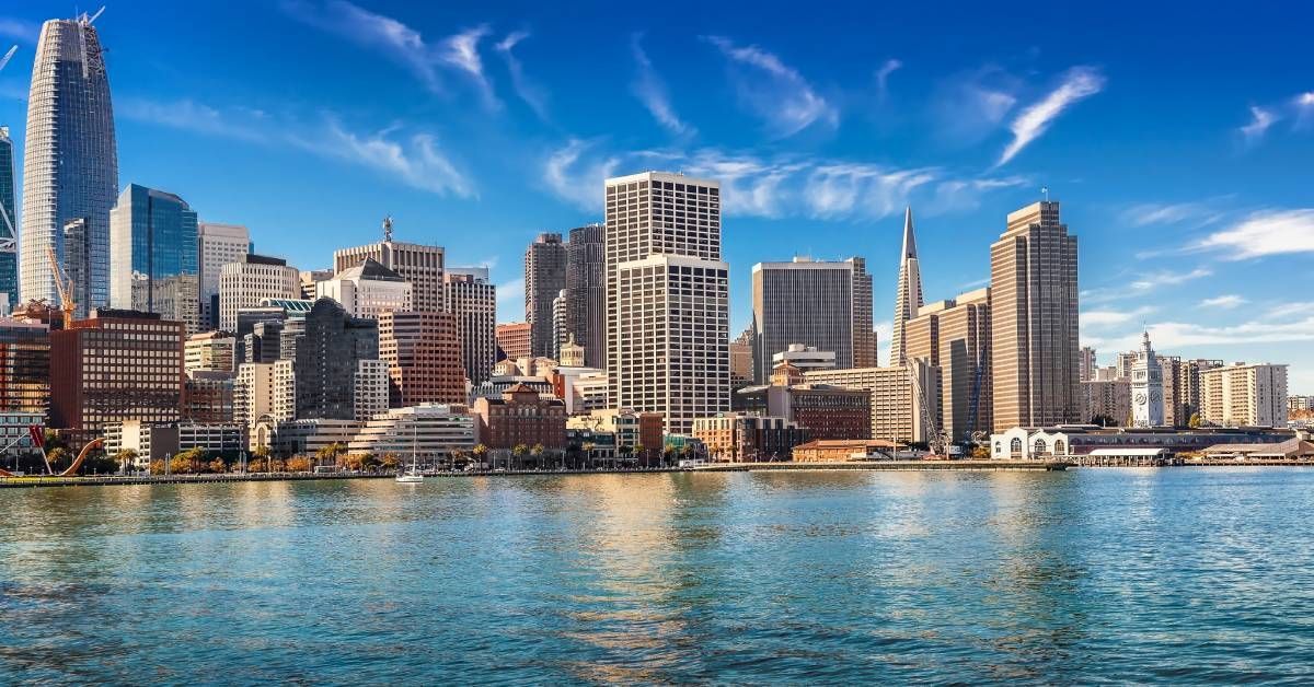 The San Francisco skyline from the other side of a bright blue bay on a clear sunny day with wispy clouds.