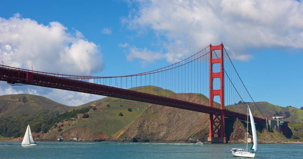 The Golden Gate Bridge stands over the water with sailboats on a bright day. There are the hills in the background.