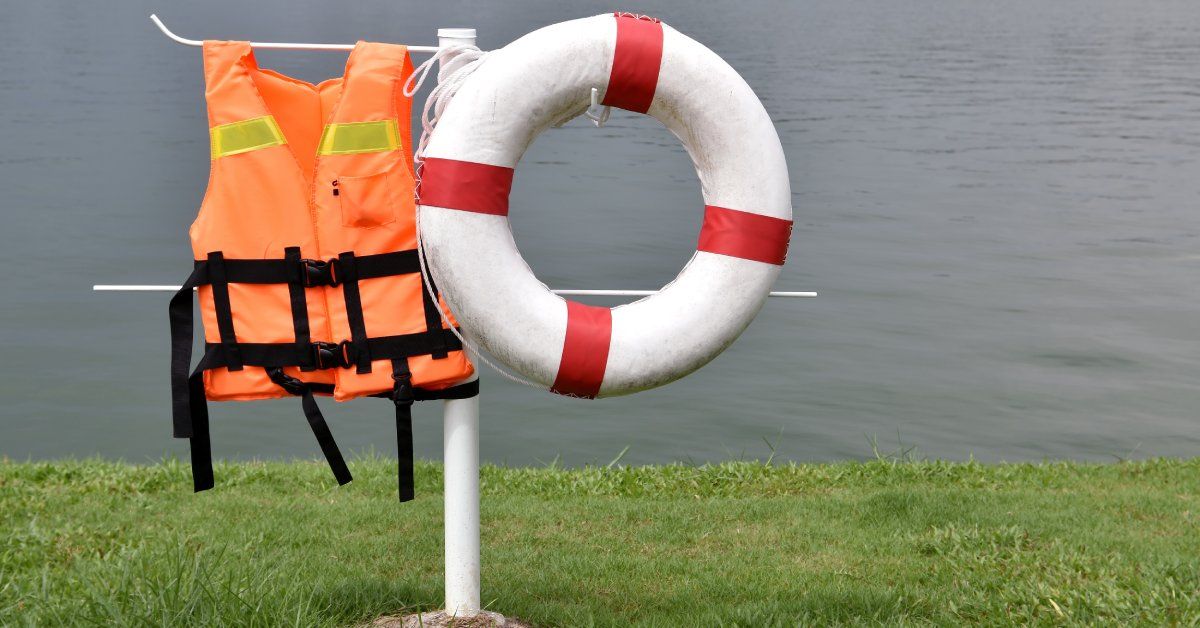 A white post in the ground with a body of water in the distance. An orange life vest and lifebuoy hang from the post.