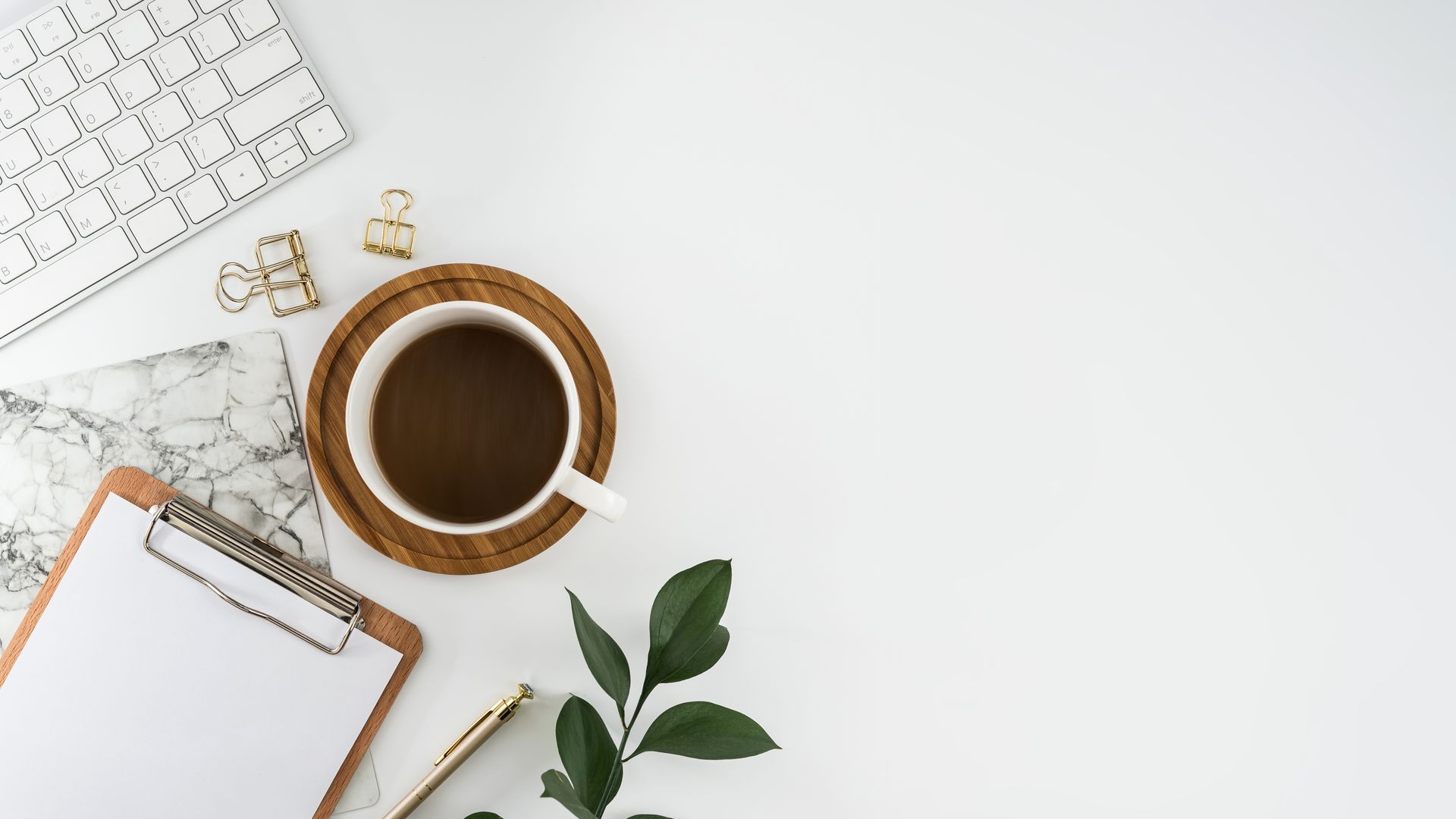 White desk with keyboard, coffee cup, clipboard, pen, leaves, and paper clips.