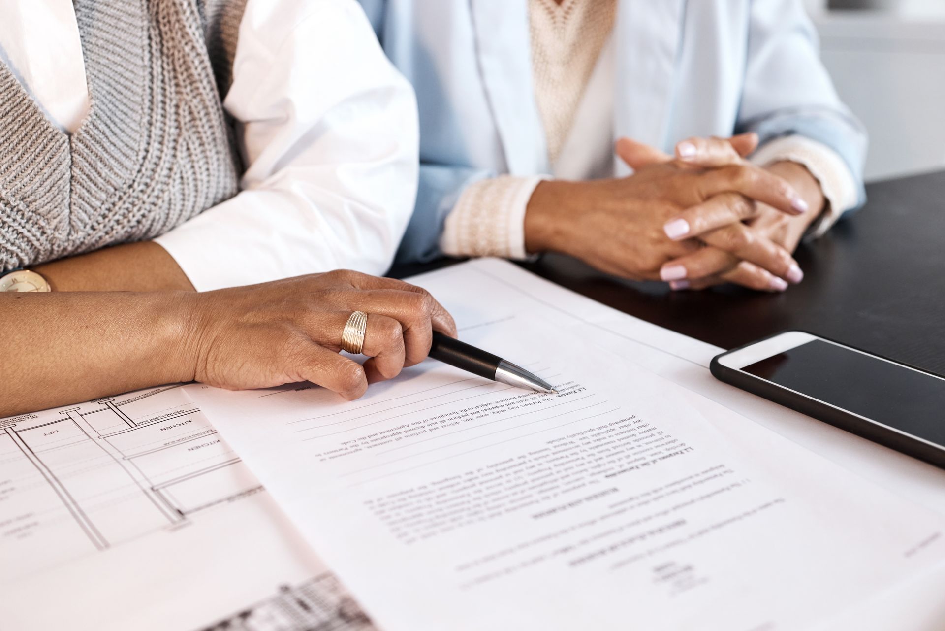 Hands pointing at documents on a table. One hand holds a pen; another rests with clasped fingers. A phone sits nearby.