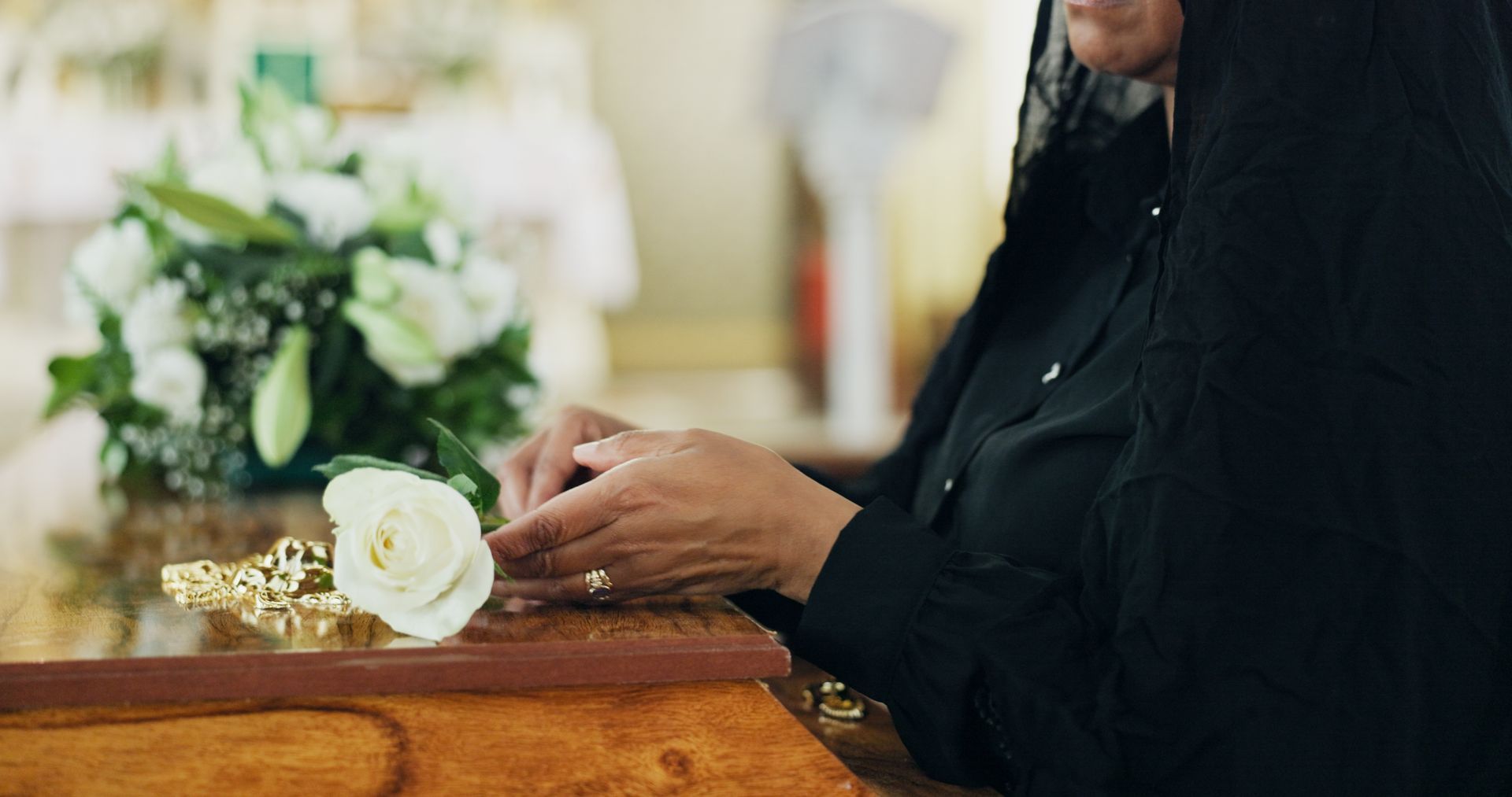 Woman in black veil places a white rose on a casket with flowers. Church setting.