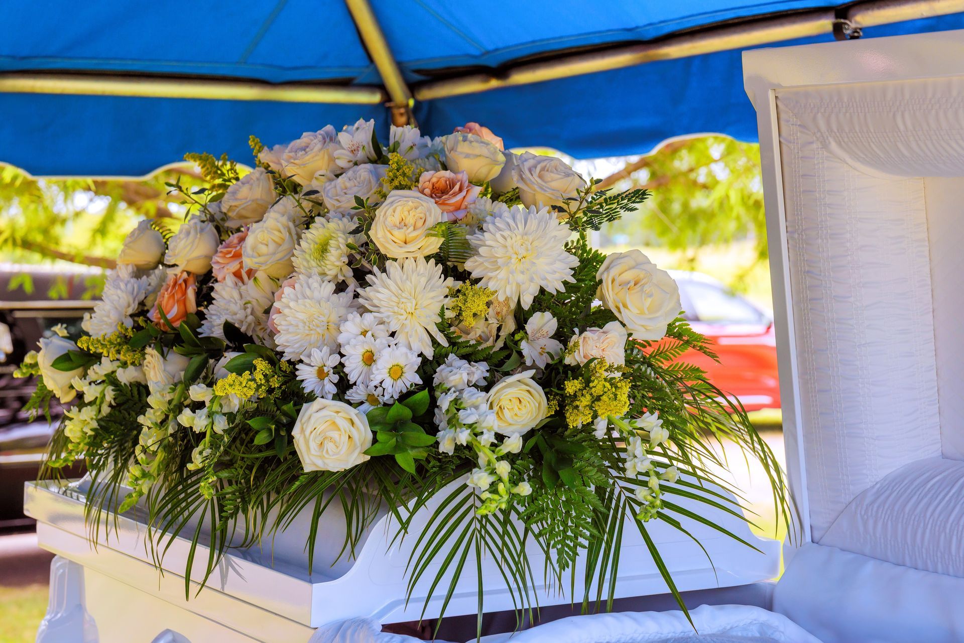 White casket with flower arrangement. Roses, daisies, and chrysanthemums atop, under a blue canopy.