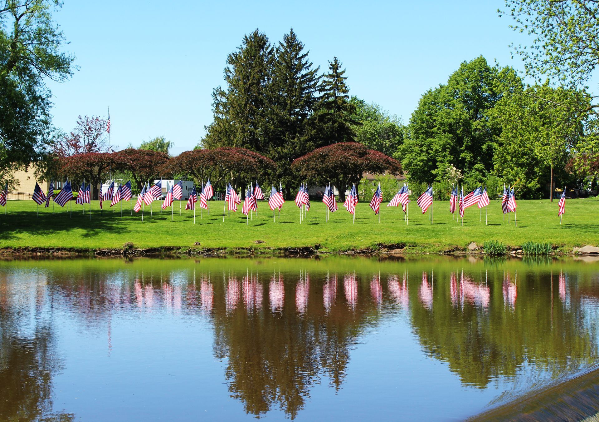 American flags line a grassy bank, reflected in the calm water of a pond, under a bright blue sky.