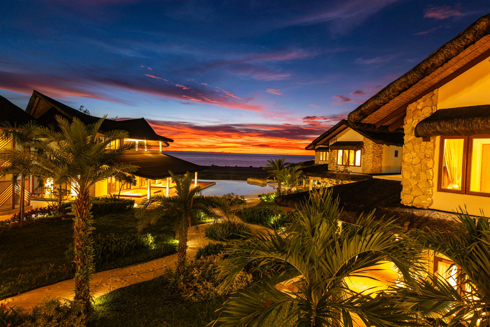 Resort villas lit at dusk beside a tropical garden and ocean under a colorful sunset sky