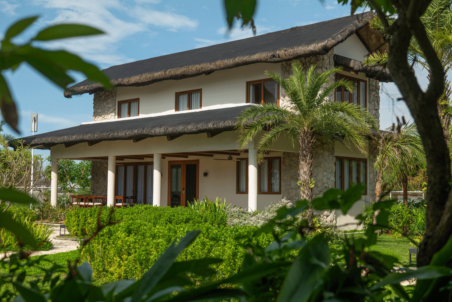 Two-story tropical house with white walls, brown roof, and lush green garden framed by palm trees
