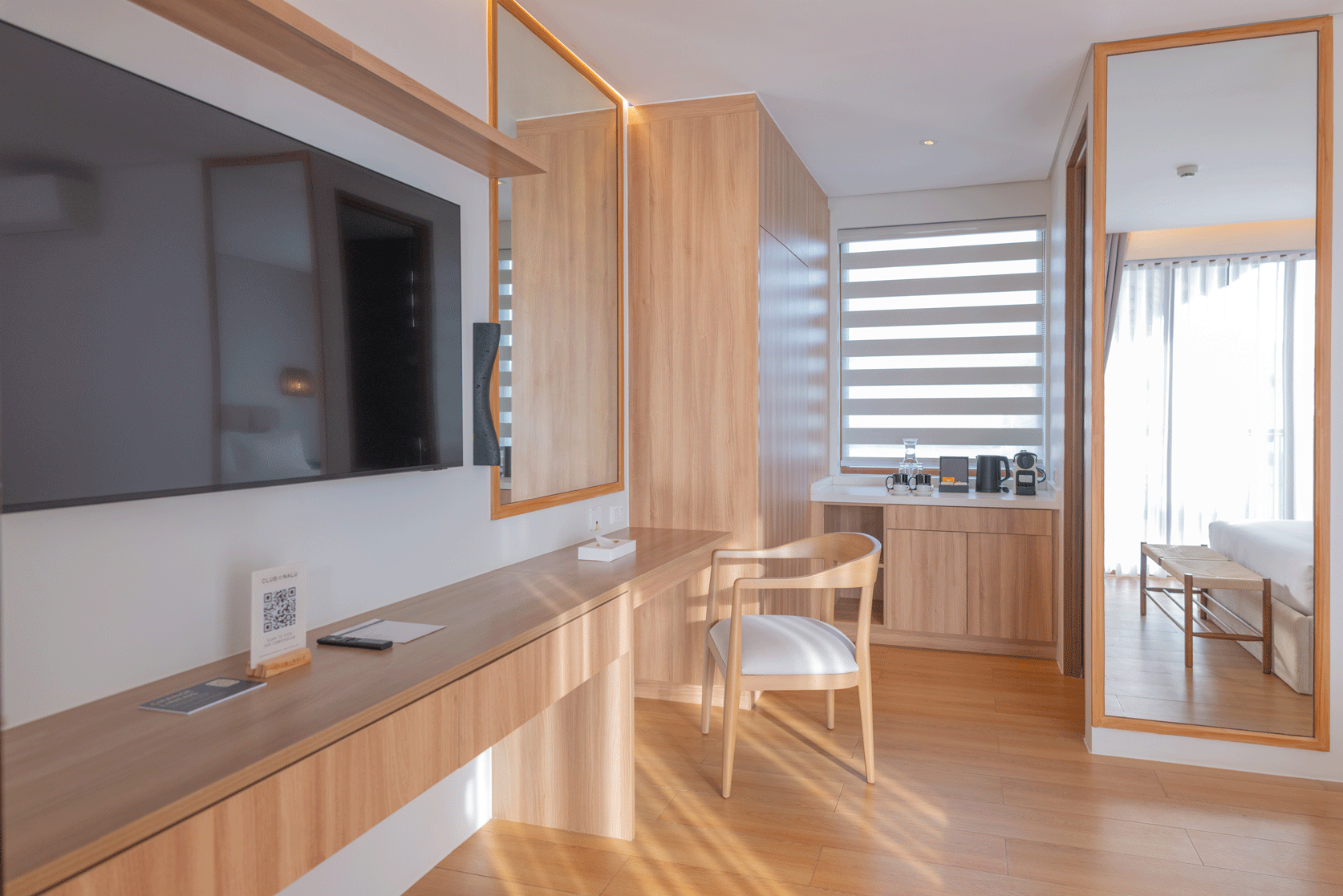 Modern minimalist room with wooden desk, chair, wall-mounted TV, and floor-to-ceiling mirror by a window.