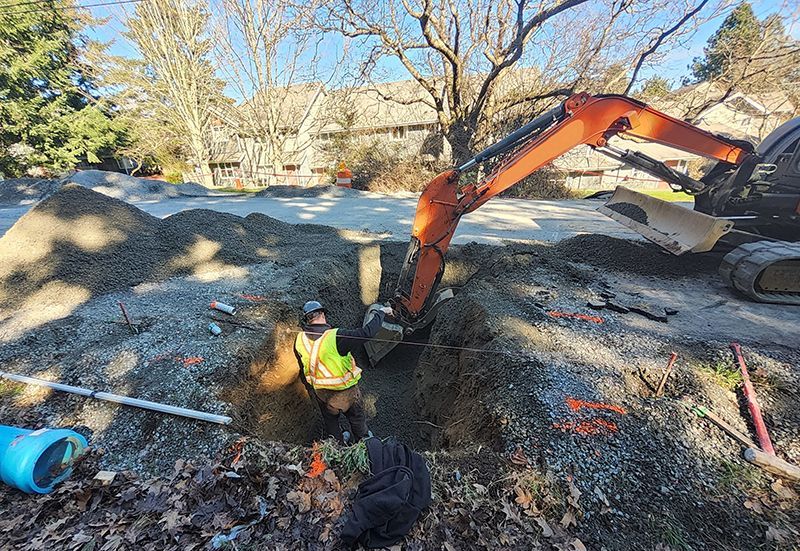 Worker in a trench, operating an excavator, near gravel piles, blue pipe, and trees on a sunny day.