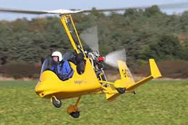 Yellow autogyro in flight over green field; pilot visible.