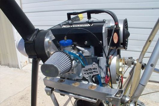 A powered paragliding engine on a stand. Black engine, propeller, blue fuel cap, and air filter.