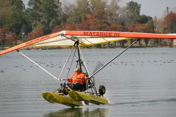 A person piloting a Maverick III ultralight aircraft with floats on a lake. Orange wing, fall foliage in background.