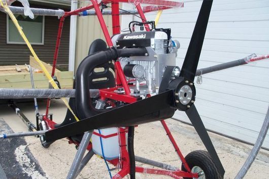 Red, open-frame ultralight aircraft with black propeller and Kawasaki engine, parked outdoors.