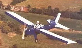 Blue and white ultralight aircraft flying over a green field, pilot visible in cockpit.