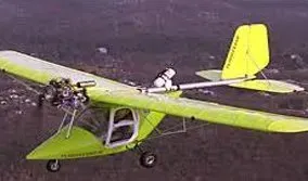 Yellow ultralight airplane in flight, pilot visible, with propeller, wings and tail.
