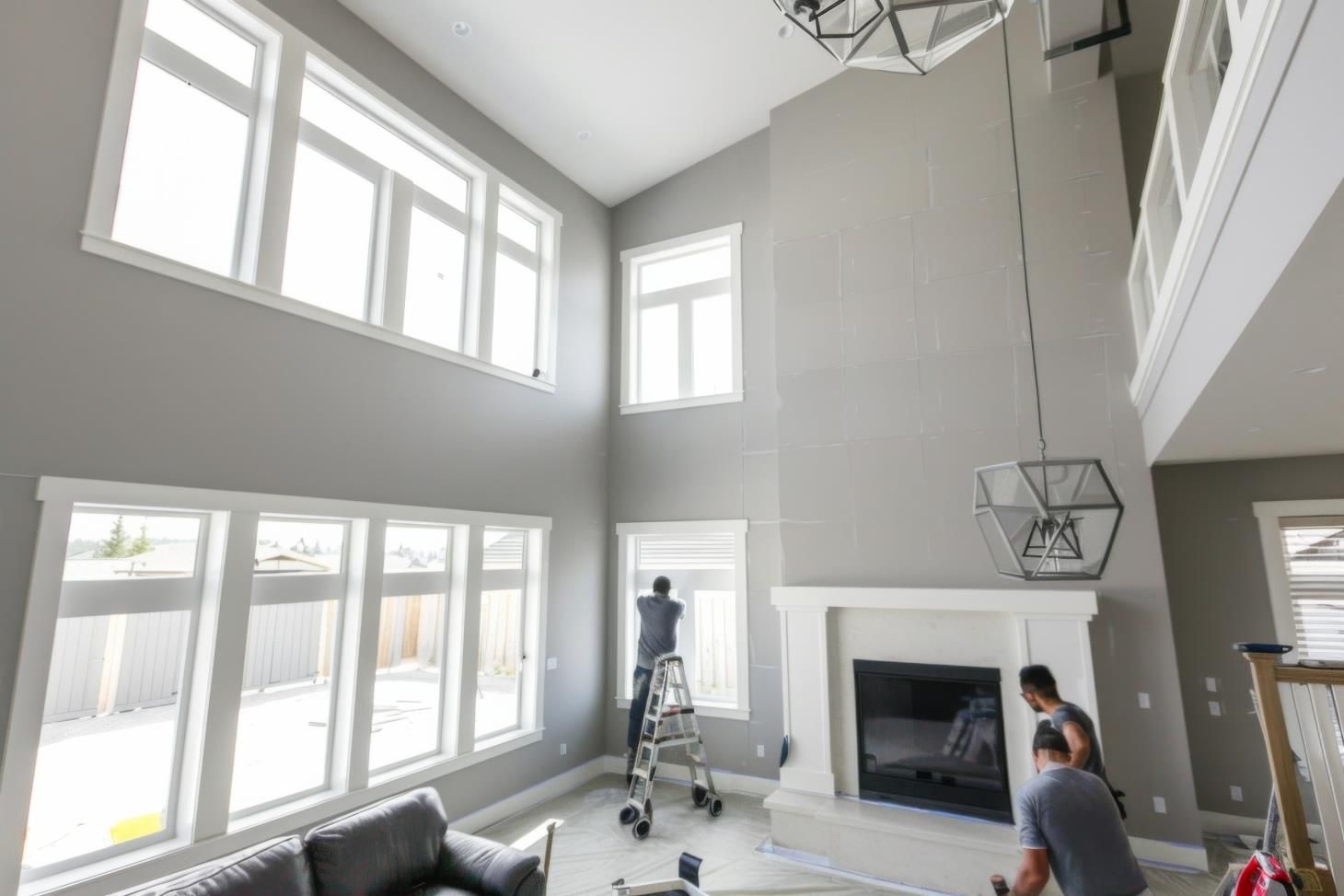 Workers painting large windows in a high-ceiling living room with gray walls and a fireplace.