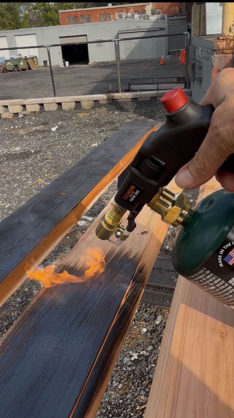 Hand holding a blowtorch flame over a black metal grate beside a wooden surface outdoors
