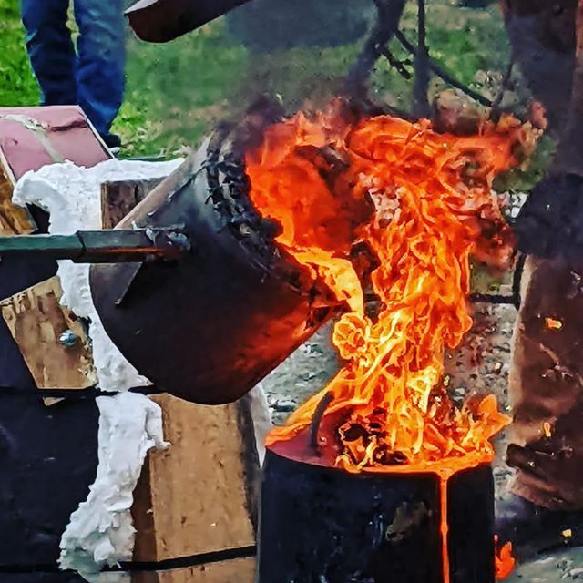 Molten metal being poured from a fiery crucible into a mold, outdoors. Orange flames and hot liquid metal.