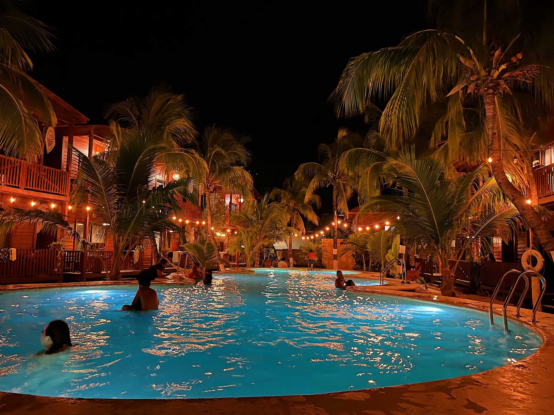 Piscina nocturna de un complejo turístico con luces brillantes, palmeras y gente nadando bajo el cielo oscuro.