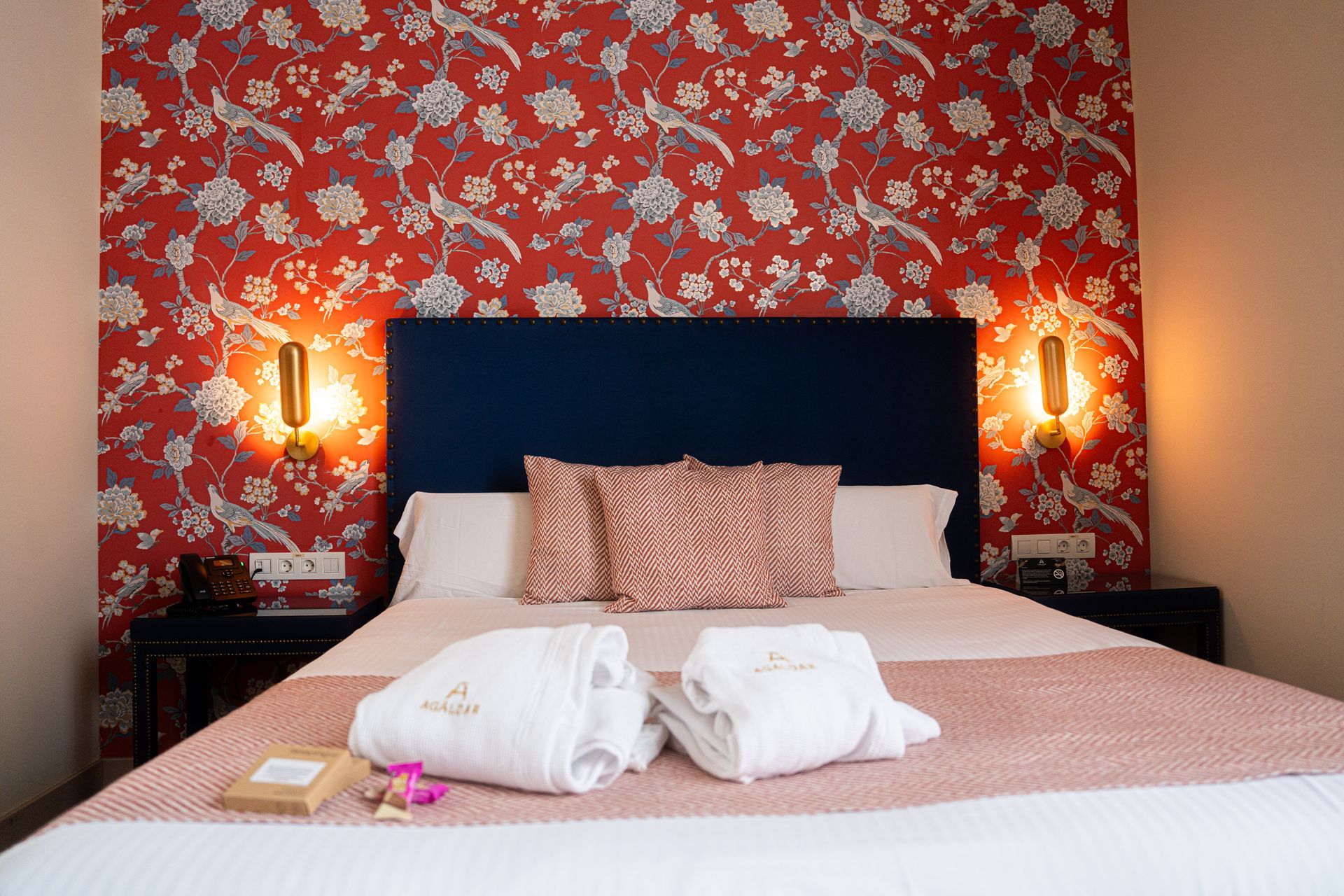 Cama de hotel con ropa de cama rosa, papel pintado floral rojo, cabecero azul marino y lámparas de noche.