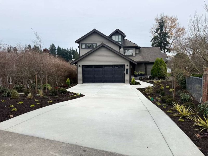 Grey house with black garage door, long concrete driveway, and landscaped yard.