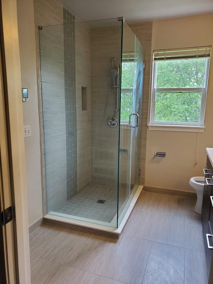 Modern bathroom with glass shower enclosure, tan tile, and a window overlooking greenery.