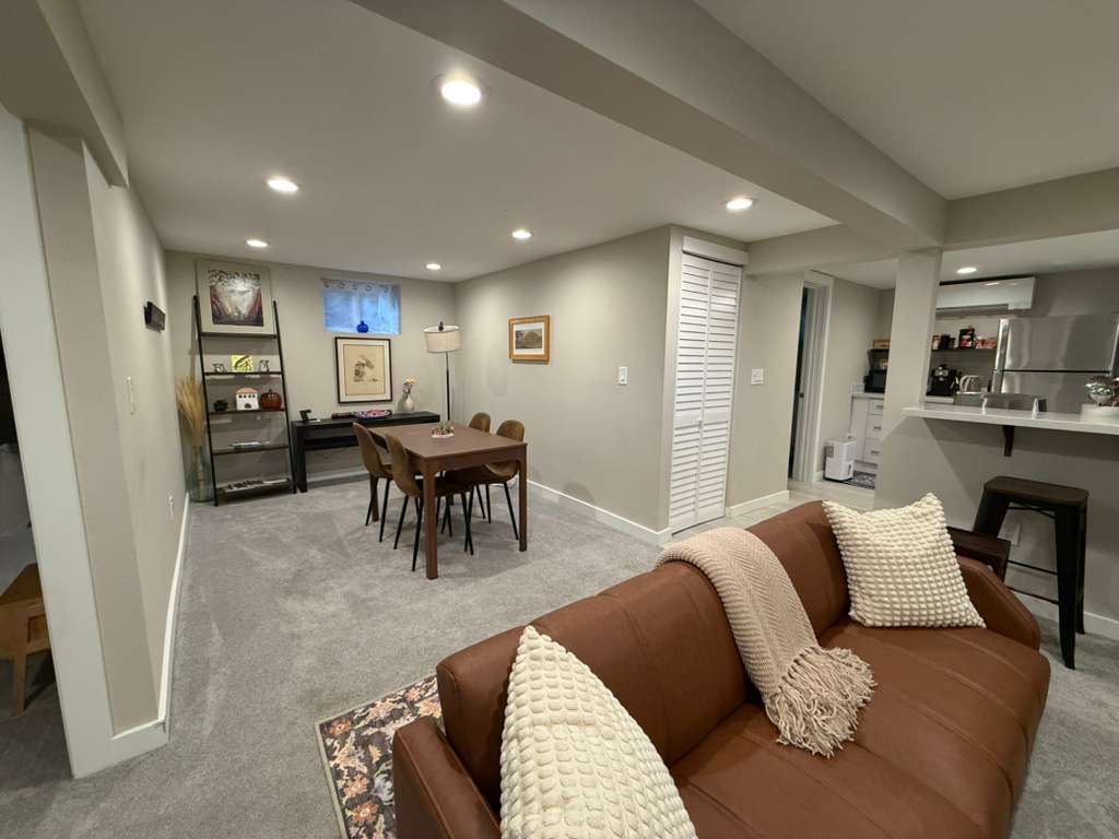 A finished basement living space featuring a brown leather sofa, dining table, and bar area. Gray carpet and walls.