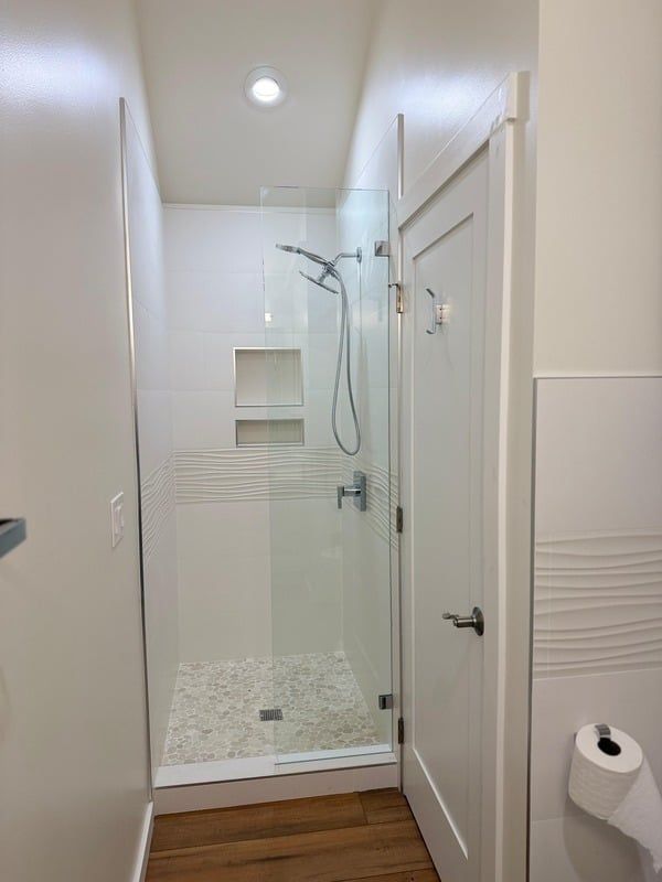 White tiled shower stall with glass door, pebble floor, and white door to the side.