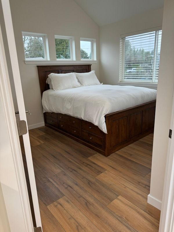 Bedroom with wood bed frame and storage drawers, white bedding, windows, and light wood floors.