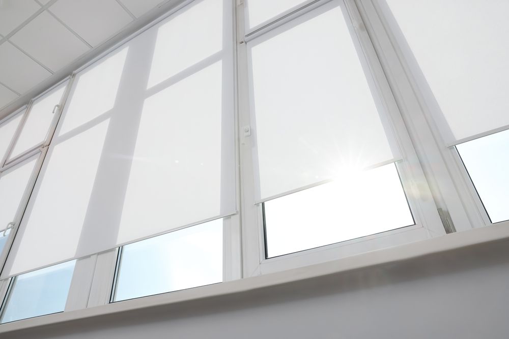White roller blinds on a window, partially covering the glass, with bright sunlight visible.