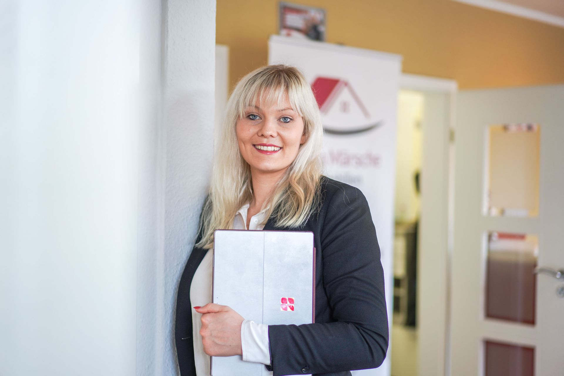 Eine Frau im Blazer hält ein Dokument in der Hand und lächelt. Sie steht in einem Büro.