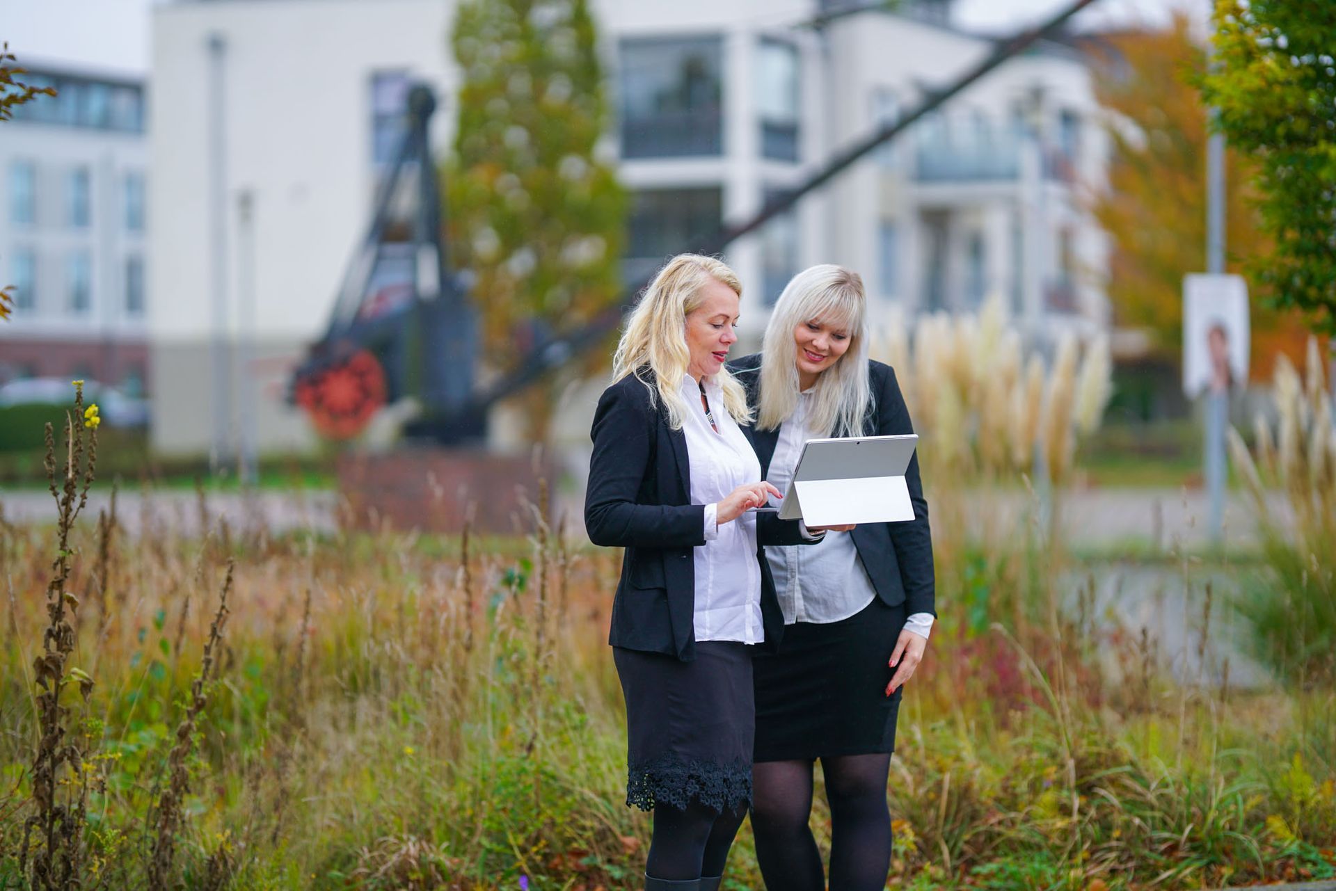 Zwei Frauen in Geschäftskleidung stehen im Freien und schauen lächelnd auf ein Tablet.