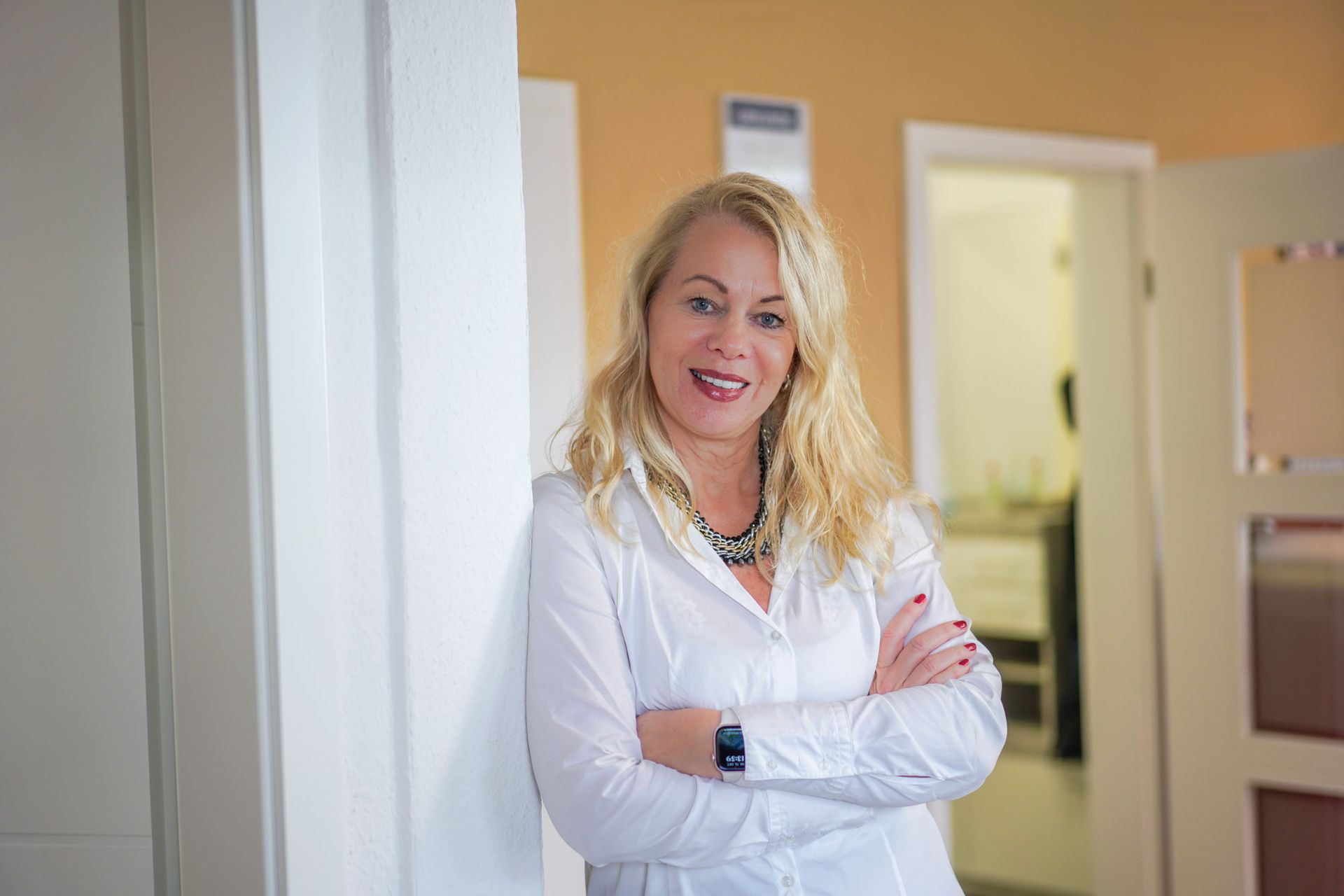 Portrait einer Immobilienmaklerin, die lächelnd mit verschränkten Armen an einer weißen Wand steht.