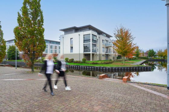 Zwei verschwommene Gestalten gehen an modernen weißen Gebäuden und einem von Bäumen gesäumten Kanal vorbei.