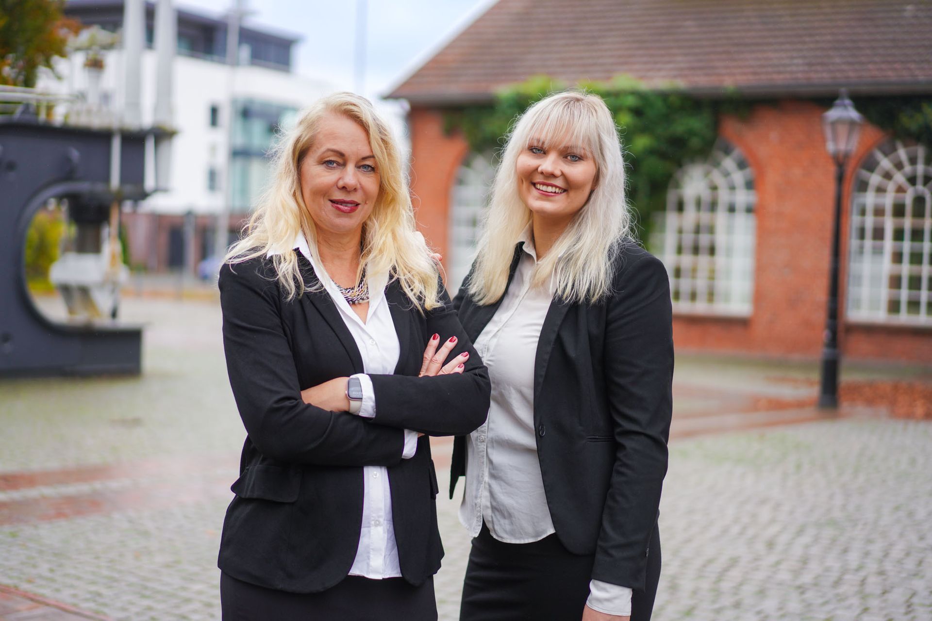 Zwei Frauen in schwarzen Anzügen stehen mit verschränkten Armen im Freien und lächeln. Im Hintergrund ein rotes Backsteingebäude.