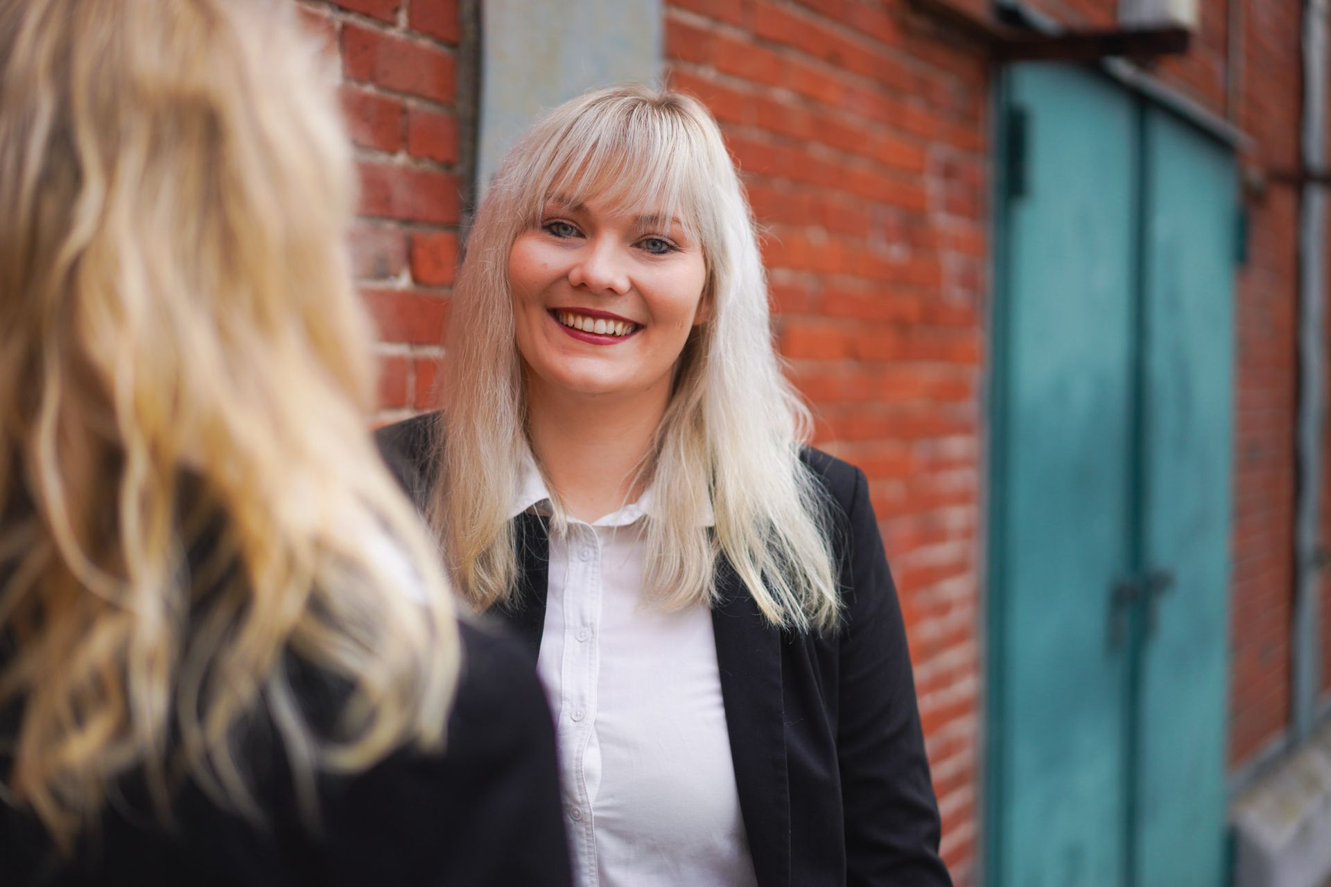 Eine blonde Frau im schwarzen Blazer lächelt vor einer Backsteinmauer, während im Vordergrund eine weitere Person unscharf zu sehen ist.