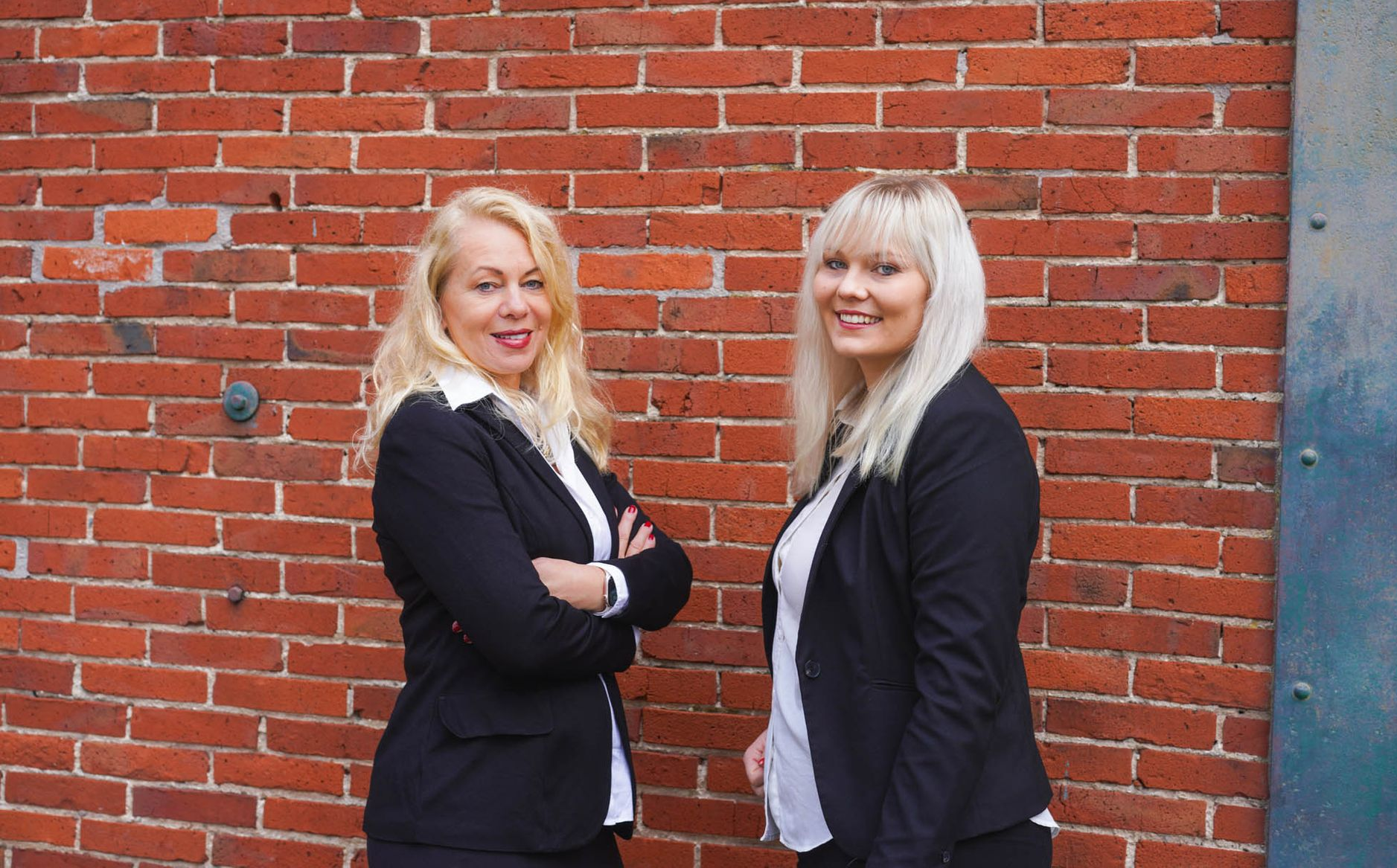 Zwei Frauen posieren vor einer Backsteinmauer und lächeln in die Kamera.