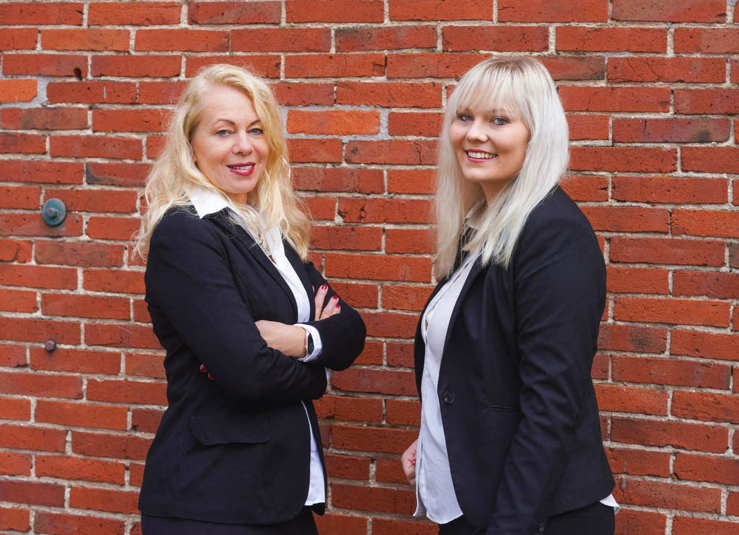 Zwei Frauen in Businessanzügen stehen lächelnd vor einer Backsteinmauer.