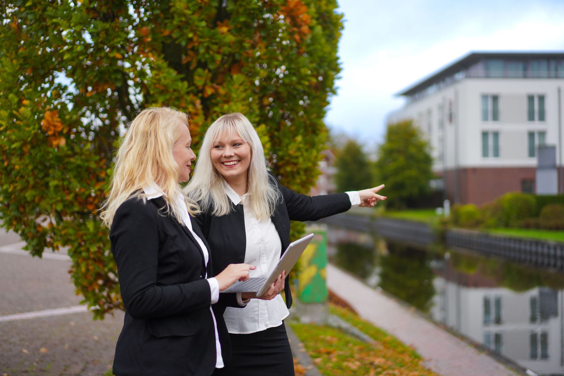 Zwei Frauen stehen lächelnd an einem Kanal. Eine zeigt in eine Richtung, die andere hält ein Laptop. Im Hintergrund ist ein Gebäude zu sehen.