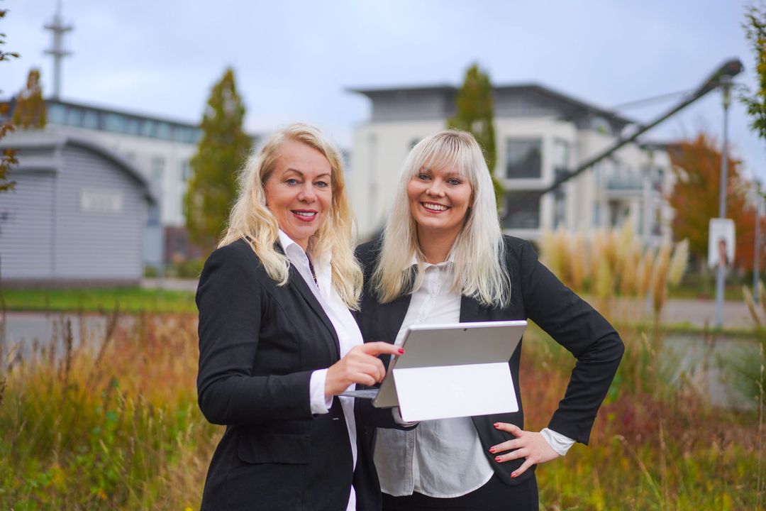 Zwei Frauen in Businessanzügen stehen im Freien, halten ein Tablet in der Hand und lächeln.