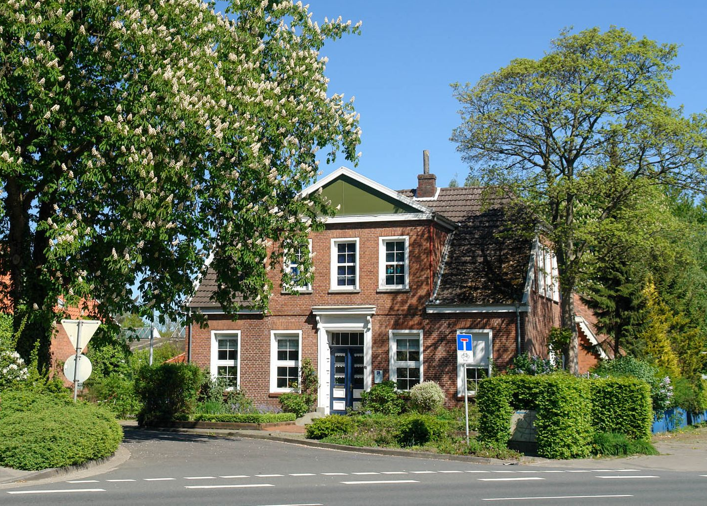 Backsteinhaus mit weißen Zierelementen, umgeben von Bäumen und Sträuchern, an einem sonnigen Tag.