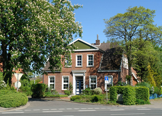 Backsteinhaus mit weißen Zierelementen, umgeben von Bäumen und Sträuchern, an einem sonnigen Tag.