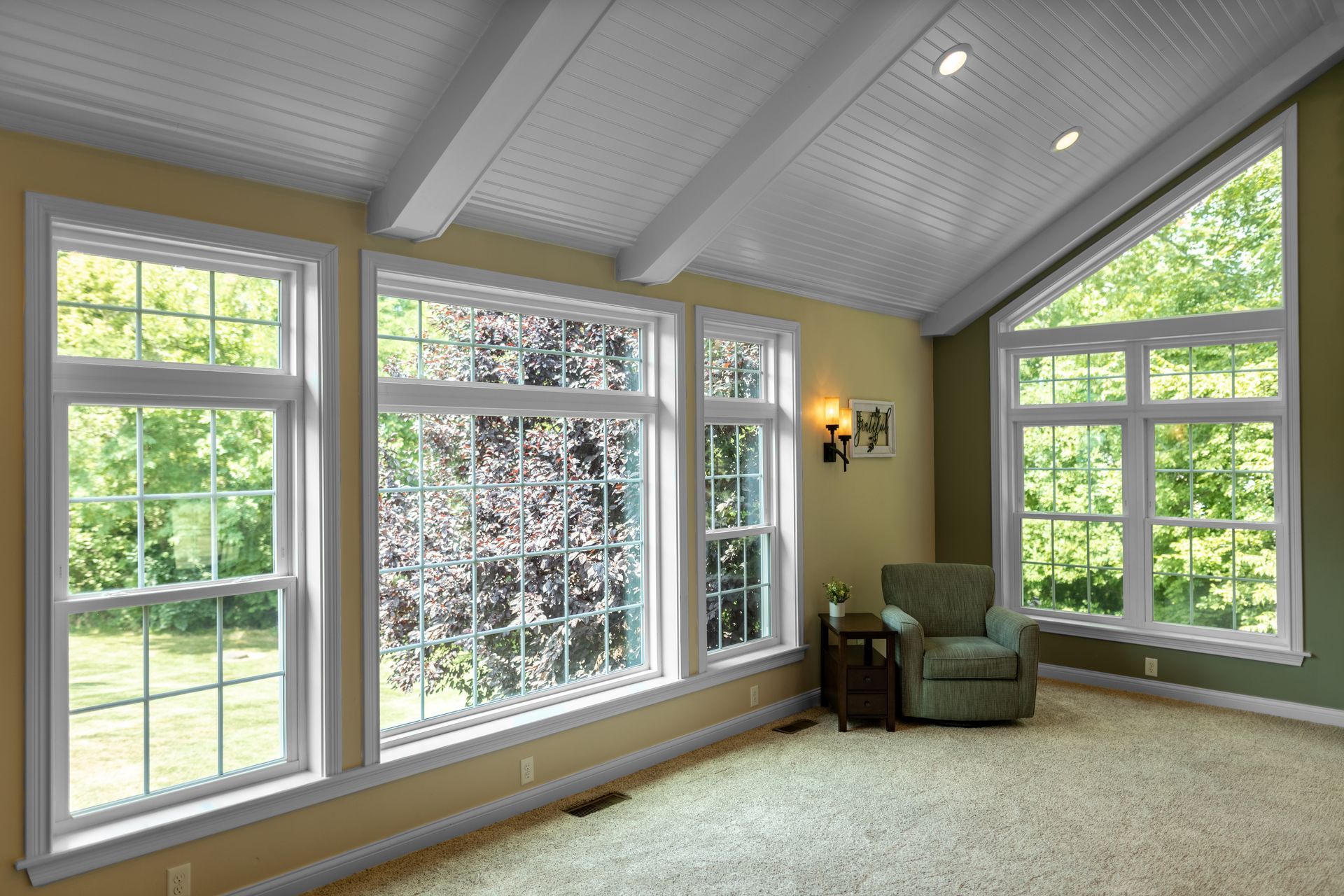 Room with multiple windows looking onto trees; light green walls, light carpet, and white ceiling beams.