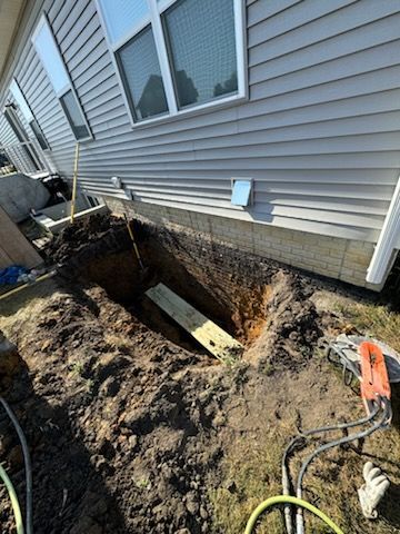 A deep excavation next to a house's foundation. Wood is placed in the hole. Tools are on the ground.