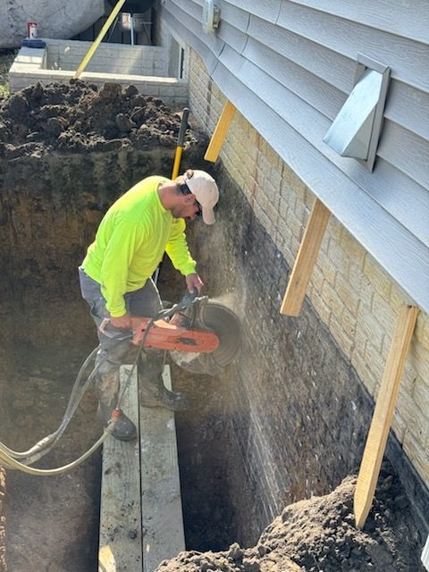 Man cutting concrete wall with a saw; dirt and building in background.