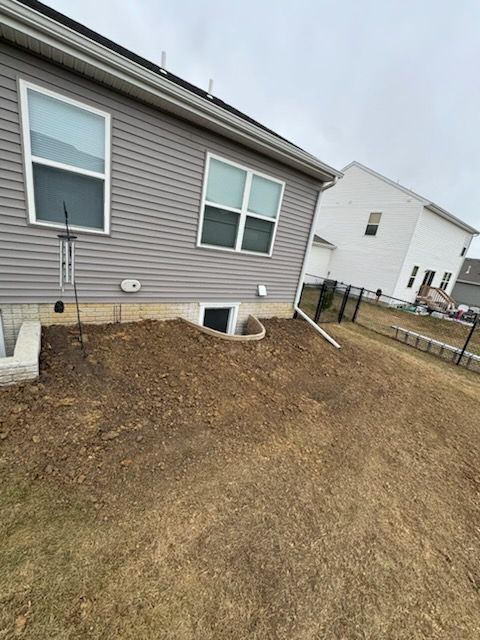 Backyard with gray siding, small window, and dirt ground. Black fence and house in background.