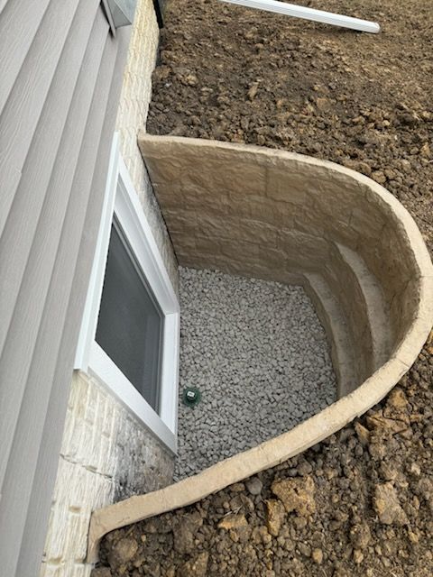 Window well with gravel base outside a building. Tan brick and tan retaining wall.