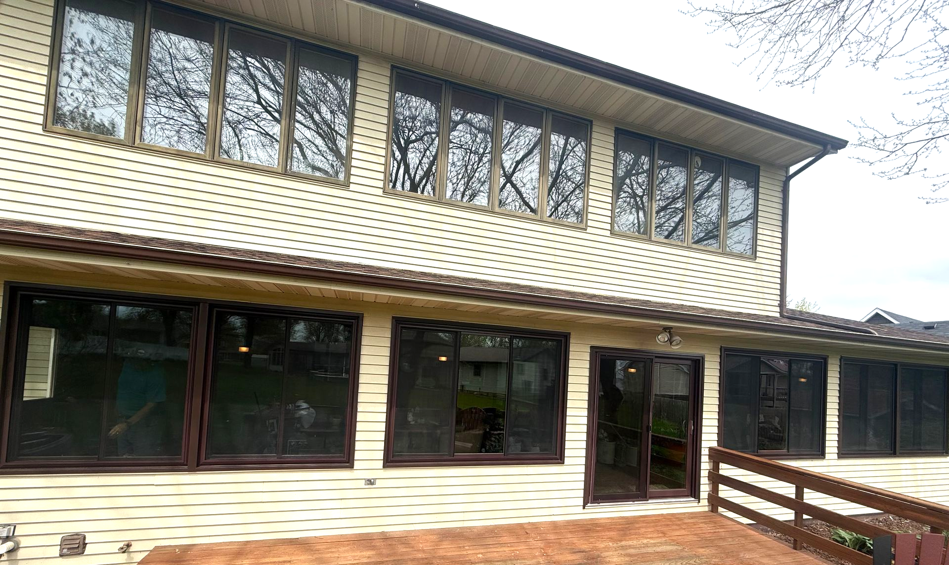 Two-story house with tan siding, brown trim, and large windows reflecting trees.